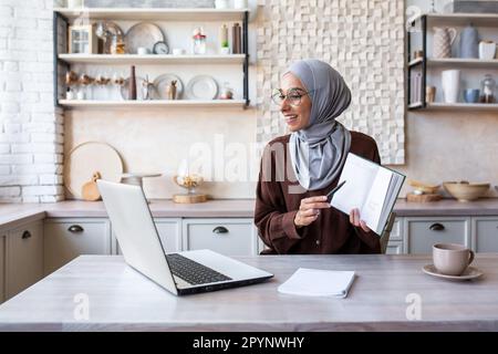 Online-Schulung. Eine junge muslimische Frau in einem Hidschab unterrichtet zu Hause per Zoom von einem Laptop aus. Zeigt der Kamera ein Buch, ein Notizbuch, erklärt, liest Stockfoto