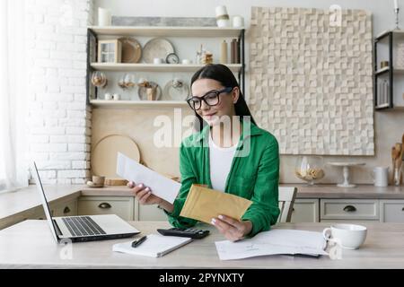 Lächelnde junge Frau, die zu Hause mit Laptop und Dokumenten arbeitet. Er öffnet den Brief aus dem Umschlag, den er erhalten hat. Ich habe gute Neuigkeiten. Stockfoto