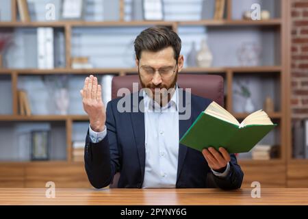 Porträt eines jungen Mannes im Anzug, eines Pastors. Ein Priester saß im Büro am Tisch, hielt ein heiliges Buch in seinen Händen, verbeugte sich und betete, predigte online, aus der Ferne/ Stockfoto