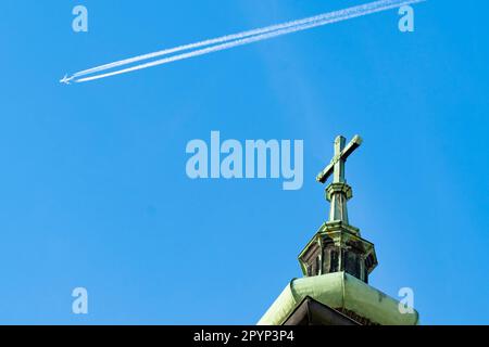 Barocker Kirchturm mit einer grünen Patina mit einem Kreuz im Hintergrund und einem Flugzeug, das einen Kontrast zieht Stockfoto