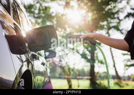 Junge Frau, die das Ladegerät in einem schwarzen Elektroauto einsteckt, Konzept für erneuerbare Energien Stockfoto