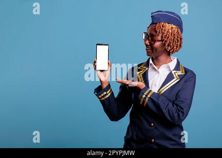 Weibliche Flugbegleiterin in Uniform hält Handy mit leerem Bildschirm für Werbung isoliert auf blauem Hintergrund. Professionelle afroamerikanische Stewardess, die in Studioaufnahmen in die Kamera schaut. Stockfoto