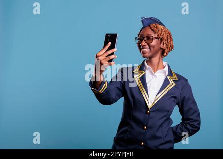 Lächelnde weibliche Flugbegleiterin in Uniform mit Videogespräch mit Handy. afroamerikanische Stewardess, die mit einem tragbaren Gerät spricht, im Studio, aufgenommen vor blauem Hintergrund. Stockfoto