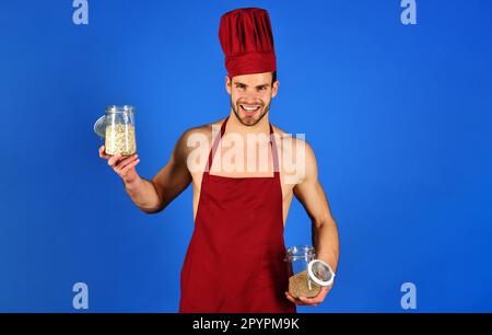 Ein lächelnder männlicher Koch mit burgunderfarbenem Hut und Schürze hält Glasgefäße mit Grobgrieß und Müsli. Professioneller Koch mit Kochhut. Gesundes Frühstück. Diät, Grütze Stockfoto