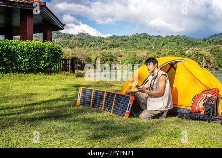 Junger Reisender, entspannen Sie sich und genießen Sie das Camping. Handstecker in tragbarem Solarpanel-Lade-Smartphone über Mehrzweckkabel, Stockfoto