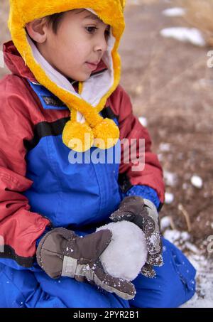 Ein nachdenklicher lateinischer kleiner Junge kniet im Schnee mit einem Schneeball in den Händen, trägt einen Schneeanzug, einen gelben Hut und Handschuhe. Stockfoto