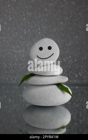 Steine mit gezogenem fröhlichem Gesicht und grünen Blättern auf grauem Hintergrund. Zen-Konzept Stockfoto