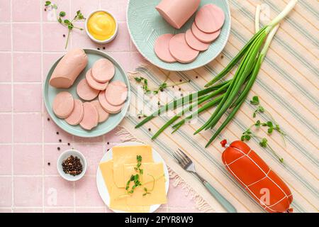 Zusammensetzung mit gekochter Wurst auf dem Teller, grünen Zwiebeln und Sauce auf einem rosa Fliesentisch Stockfoto