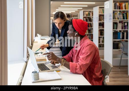 Ein fokussiertes, multiethnisches Studentenpaar spricht über die Prüfung eines Studienprojekts mit einem Laptop in der Universitätsbibliothek Stockfoto