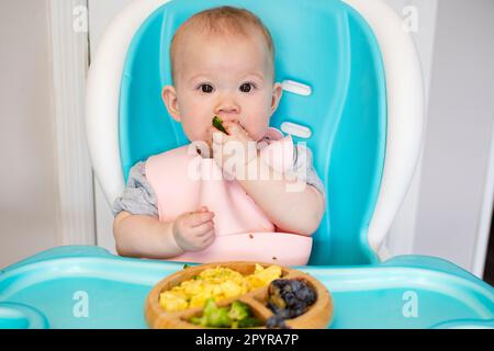 Baby isst Brokkoli. Säuglingsprothetik. Entwöhnung. Gesunde Ernährung. Kaukasisches Mädchen, das in einem Hochstuhl sitzt und ihr Mittagessen isst Stockfoto