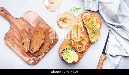 Gourmet-Mittagessen mit Avocado-Lachs-Toasts auf Roggenbrot und Rosenweingläsern, grauer Tischhintergrund, Blick von oben Stockfoto