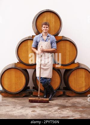 Porträt, Besen und Eichenfässer mit einem Mann, der den Weinkeller einer Getränkedestillerie reinigt. Selbstbewusst, Schürze und männlicher Reiniger in einem Weingut für die Stockfoto