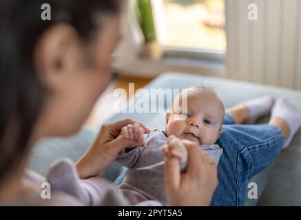 Porträt eines süßen, zufriedenen Neugeborenen, das zu Hause auf den Beinen der Mutter auf dem Sofa liegt, Spaß hat und spielt. Mutterschaftsliebe, Pflege und Freude Konzept Stockfoto