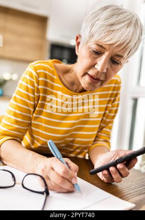 Reife Frau, die ihre Haushaltsrechnungen mit dem Handy berechnet und zu Hause Kosten und Einsparungen am Esstisch schreibt Stockfoto