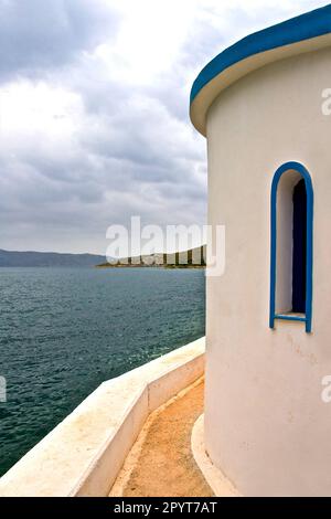 Kleine Kapelle mit weißen Dächern auf Klippe über Meer und Kleine Bucht unter einem dramatischen Himmel auf einer griechischen Insel Stockfoto