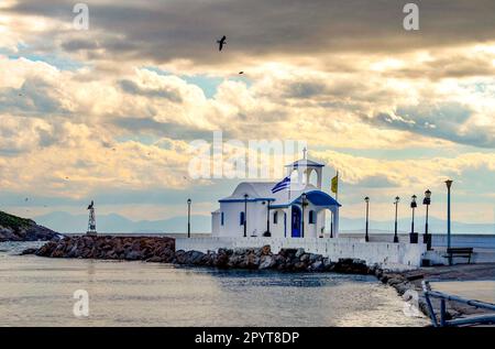 Kleine Kapelle mit weißen Dächern auf Klippe über Meer und Kleine Bucht unter einem dramatischen Himmel auf einer griechischen Insel Stockfoto