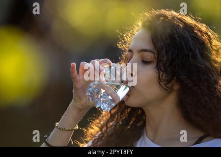 Hübsche junge Frau, die im Park Wasser aus Glas trinkt Stockfoto