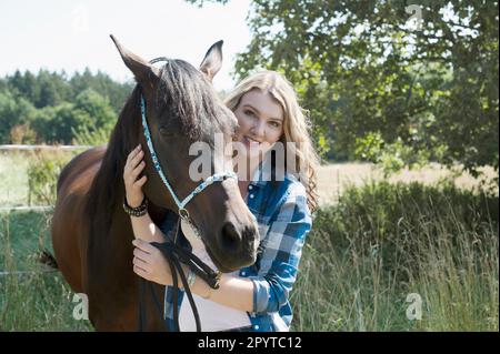 Junge Frau, die ihr braunes Pferd streichelte, auf einer Wiese stand und lächelte, Bayern, Deutschland Stockfoto