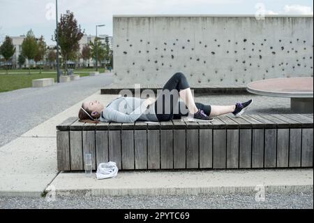 Junge Frau, die eine Pause macht, nachdem sie auf einer Holzbank gelegen und Musik gehört hat, Bayern, Deutschland Stockfoto