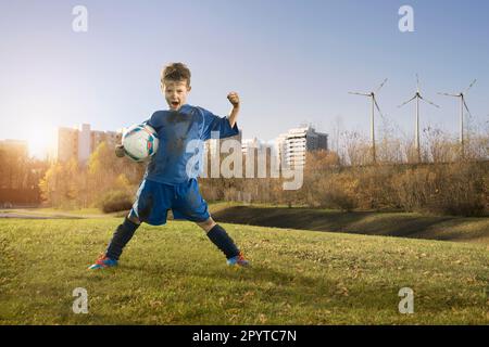 Dreckiger Fußballspieler, der auf dem Feld und den Windturbinen jubelt, mit Stadt im Hintergrund, Bayern, Deutschland Stockfoto