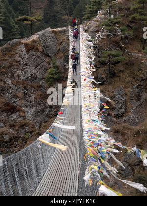 Buddhistische Gebetsflaggen, die an eine metallische Hängebrücke namens „Hillary Bridge“ gebunden sind, und Wanderungen im Hintergrund bei der Wanderung im Everest Base Camp. Stockfoto