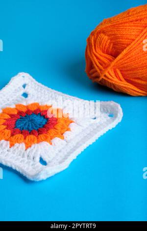 Granny Square mit Häkeldesign und weißem Quadrat mit orangefarbenem, rotem und blauem Muster in der Mitte und orangefarbenem Garnknäuel auf blauem Hintergrund. Stockfoto