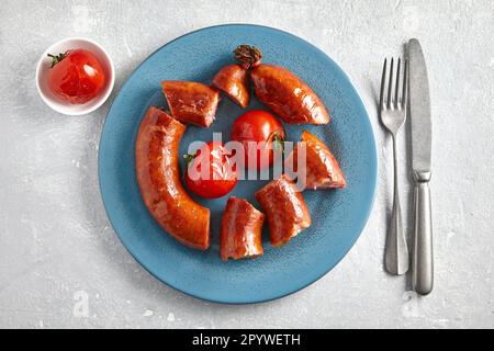 Gebratene Schweinewurst in Stücke geschnitten auf einem blauen Teller mit Tomaten und Besteck auf hellgrauem Steinhintergrund. Draufsicht mit Kopierbereich Stockfoto