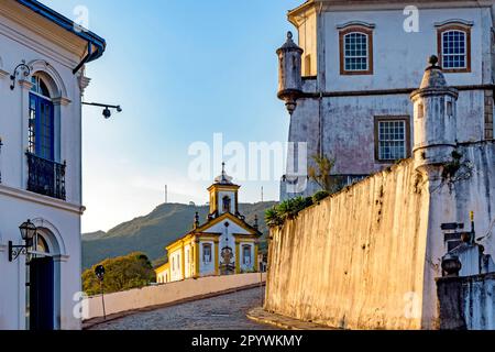 Ankunft im historischen Zentrum von Ouro Preto mit Häusern im Kolonialstil, Denkmälern, Kirchen und alten Gebäuden mit Hügeln im Hintergrund bei Sonnenuntergang Stockfoto