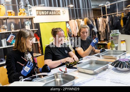 05. Mai 2023, Baden-Württemberg, Freiburg: Besucher Ira Pel (l) und ein Medienvertreter (r) lassen Karen Graf (M), Umwelterzieherin bei Abfallwirtschaft und Stadtreinigung Freiburg GmbH (ASF), ihnen zeigen, wie man einen Weinflaschenhalter für die Wand aus einem alten Rekord herstellt, während eines Upcycling-Workshops auf der Greenflair-Nachhaltigkeitsmesse. Im nächsten Jahr soll der neue Second-Hand-Laden Fundfabrik der ASF eröffnet werden. Das Konzept ist „einzigartig“, neben einer ausgewählten Auswahl an gebrauchten Möbeln, Möbeln, Haushaltswaren und gebrauchter Kleidung bietet die Fundfabrik ihren Gästen auch „Upcycll“ Stockfoto