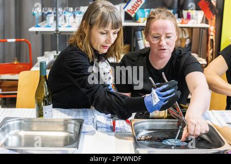 05. Mai 2023, Baden-Württemberg, Freiburg: Besucher Ira Pel (l) zeigt Karen Graf (r), Umwelterzieherin der Abfallwirtschaft und Stadtreinigung Freiburg GmbH (ASF), wie man einen Weinflaschenhalter für die Wand aus einem alten Rekord herstellt, während eines Upcycling-Workshops auf der Nachhaltigkeitsmesse „Greenflair“. Im nächsten Jahr soll der neue Second-Hand-Laden Fundfabrik der ASF eröffnet werden. Das Konzept ist „einzigartig“, neben einer ausgewählten Auswahl an gebrauchten Möbeln, Möbeln, Haushaltswaren und gebrauchter Kleidung bietet die Fundfabrik ihren Gästen auch „Upcycling Workshops“, ein „Reparaturwerk“ Stockfoto