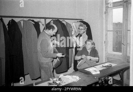 Ein Schneider untersucht die Nähte einer Jacke. Die sitzende Näherin näht das Futter eines Mantels. Unbezahltes Foto von etwa 1955 Stockfoto