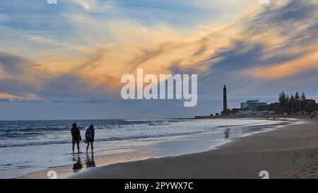 Strand, zwei Strandbesucher von hinten, Dämmerung, blau-orangefarbener Himmel, graue Wolken, Hotel, Leuchtturm, Meer, Maspalomas, Gran Canaria, Kanarische Inseln, Spanien Stockfoto