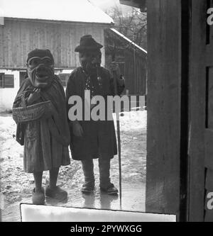 Zwei Personen mit übergroßen, handgeschnitzten Holzmasken vor ihren Gesichtern, die vor einem Haus in Oberbayern stehen (undatiertes Bild). Stockfoto