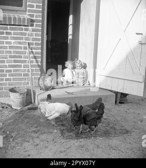 Zwei kleine Mädchen sitzen vor der Tür eines Bauernhauses und beobachten die Hühner. Unbefristetes Foto. Stockfoto