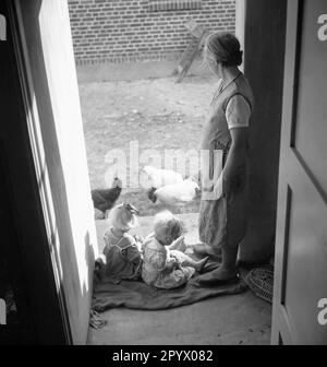 Zwei kleine Mädchen sitzen an der Schwelle eines Bauernhauses. Neben ihnen steht eine Farmerin. Alle drei sehen den Hühnern beim Essen im Hof zu. Unbefristetes Foto. Stockfoto