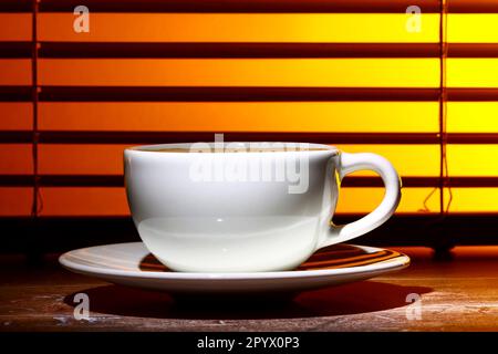 Kaffeetasse gefüllt mit schaumigem Kaffee, an einem Fensterrollo Stockfoto