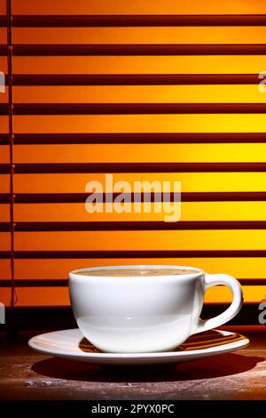 Kaffeetasse gefüllt mit schaumigem Kaffee, an einem Fensterrollo Stockfoto