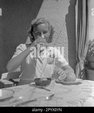 Eine junge, blonde Frau sitzt an einem Tisch und trinkt Zitronensaft aus einem Glas, vor ihr liegt ein Zitronenpresser und ein Teller mit Zitronen. Stockfoto