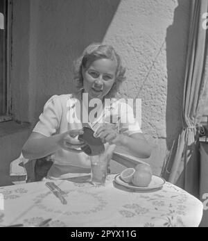 Eine junge Frau sitzt am Tisch und gießt den Saft aus einer Zitronensaftpresse in ein Glas, vor ihr stehen Zitronen auf einem Teller. Stockfoto