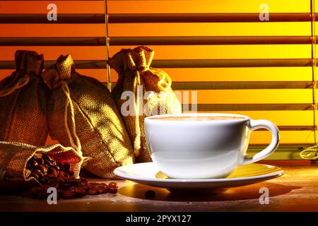 Kaffeetasse gefüllt mit Kaffee, Set vor einem Küchenfenster mit Säcken roher gerösteter Kaffeebohnen Stockfoto