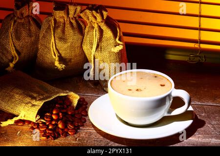 Kaffeetasse gefüllt mit Kaffee, Set vor einem Küchenfenster mit Säcken roher gerösteter Kaffeebohnen Stockfoto