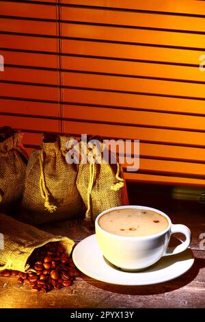 Kaffeetasse gefüllt mit Kaffee, Set vor einem Küchenfenster mit Säcken roher gerösteter Kaffeebohnen Stockfoto