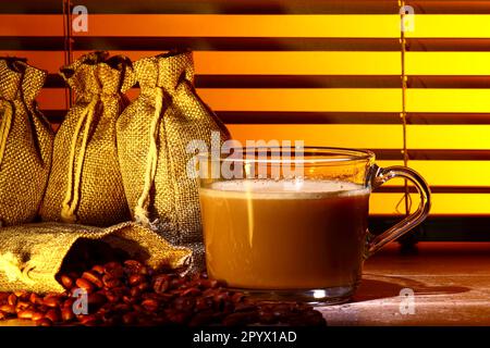 Glas-Kaffeetasse gefüllt mit Kaffee, Set vor einem Küchenfenster-Jalousien mit Säcken roher gerösteter Kaffeebohnen Stockfoto