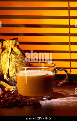 Glas-Kaffeetasse gefüllt mit Kaffee, Set vor einem Küchenfenster-Jalousien mit Säcken roher gerösteter Kaffeebohnen Stockfoto