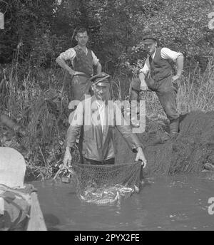 Der Jäger eines Herrenhauses präsentiert dem Fotografen seinen Fang. Das Netz ist voller Fische. Zwei weitere Männer stehen hinter ihm. Unbezahltes Foto, wahrscheinlich auf einer Farm in der Provinz Pommern in den 1930er. Stockfoto