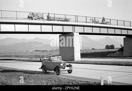 Ein Auto fährt auf der neu eröffneten Autobahn München-Salzburg. Eine Brücke führt über den Highway, über den zwei Bullock Carts fahren. Im Hintergrund die Alpen. Unbezahltes Foto, wahrscheinlich im Sommer 1939. Stockfoto