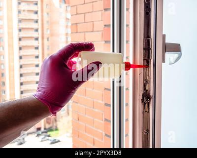 Hand in rosa Handschuh schmiert den Mechanismus des Kunststofffensterrahmens in einer Wohnung eines mehrstöckigen Stadtgebäudes aus einer Ölkanne Stockfoto