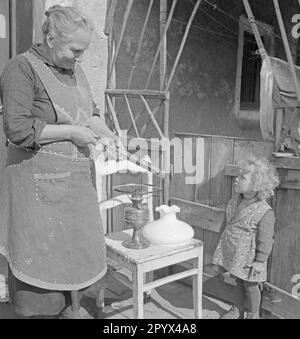 Foto einer älteren Bauernfrau in einer Schürze, die im Sommer 1936 vor einer Scheune in gut Osdorf südlich von Berlin eine Kerosinlampe mit einem neuen Docht ausrüstet. Sie wird von einem kleinen Kind beobachtet. Stockfoto