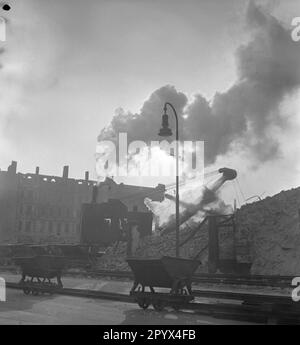 Undatiertes Foto eines Baggers, der einen Haufen Trümmer entlädt und Wagen mit Trümmer vor einem teilweise zerstörten Wohnblock lädt, bei einem Bombenanschlag in der Nähe der Ruinen der Kaiser-Wilhelm-Gedächtniskirche am Kurfürstendamm in Charlottenburg, West-Berlin, einer ehemaligen britischen Besatzungszone. Die dieselbetriebene Baumaschine nimmt Ziegel und Kies mit einem Greifer auf. Im Vordergrund, Ketten und Führungsschienen. Stockfoto