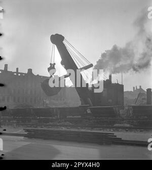 Undatiertes Foto eines Baggers, der einen Haufen Trümmer entlädt und Wagen mit Trümmer vor einem teilweise zerstörten Wohnblock lädt, bei einem Bombenanschlag in der Nähe der Ruinen der Kaiser-Wilhelm-Gedächtniskirche am Kurfürstendamm in Charlottenburg, West-Berlin, einer ehemaligen britischen Besatzungszone. Die dieselbetriebene Baumaschine nimmt Ziegel und Kies mit einem Greifer auf. Im Vordergrund, Ketten und Führungsschienen. Stockfoto